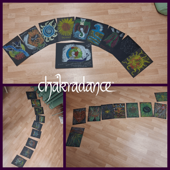 En serie av färgglada teckningar med chakra-tema som ligger på golvet, arrangerade i en båge. Flera konstverk syns tydligt, med olika mönster och symboler.